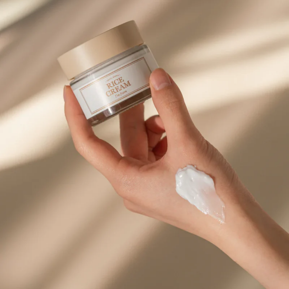 Hand holding a jar of 'Rice Cream' with a swatch on skin against a beige background