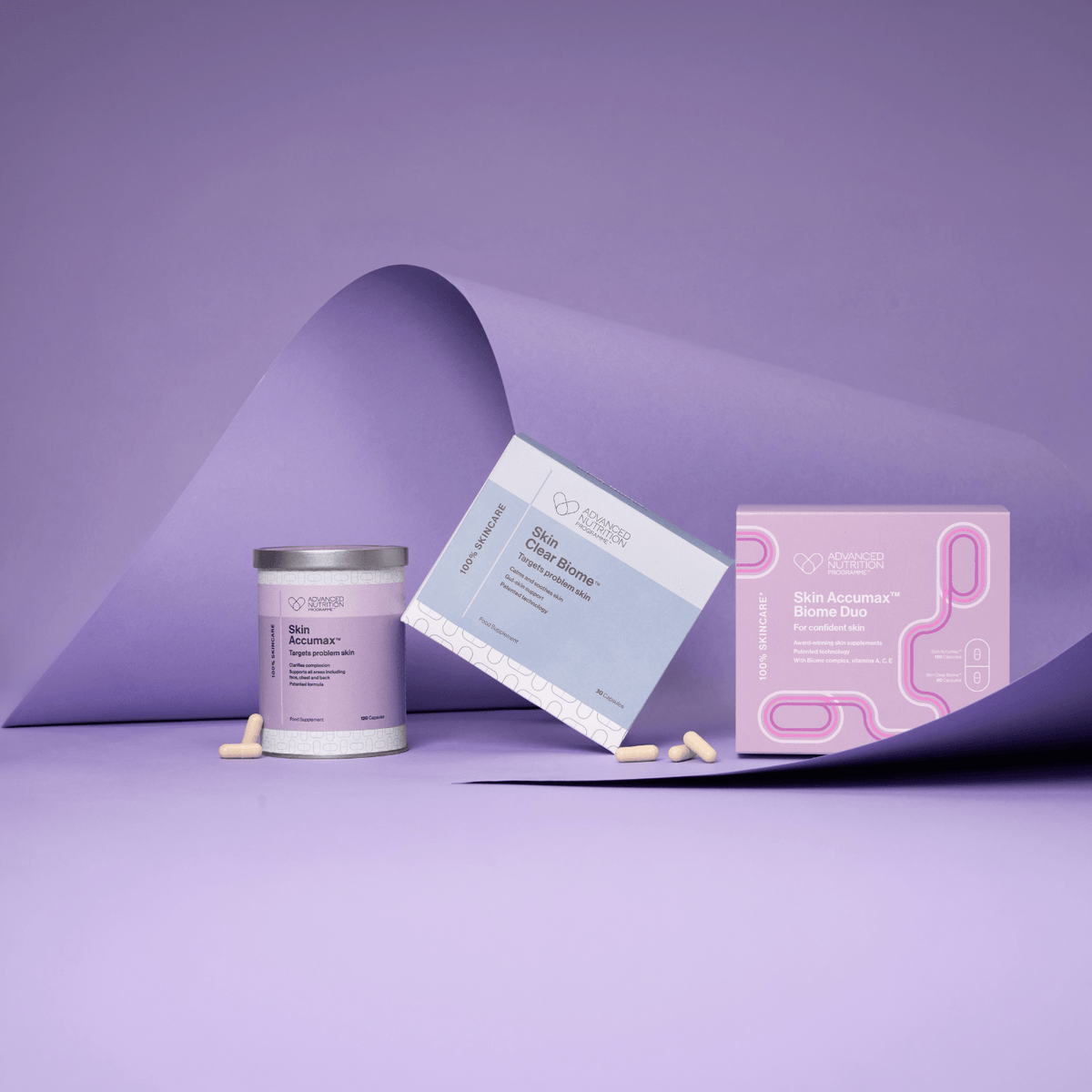 Advanced Nutrition Programme Skin Accumax Biome Duo box, Skin Accumax tin and Skin Clear Biome box arranged on curled lilac paper, with a few capsules scattered in front.